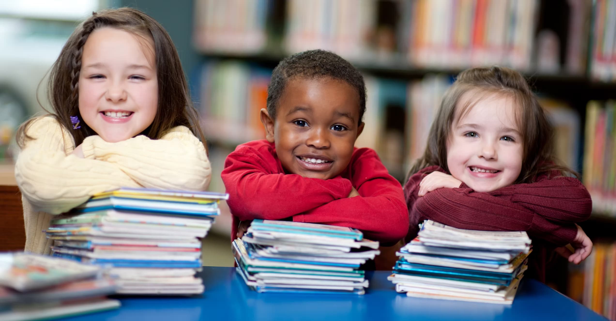 Three diverse, smiling children with books in a bright library setting - representing the families served by Muskegon Pediatrics